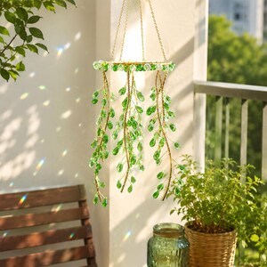 May include: A hanging mobile featuring a circular gold-toned frame suspended by three thin chains. Attached to the frame are multiple cascading strands of wire adorned with faceted, teardrop-shaped green glass crystals that catch the sunlight.