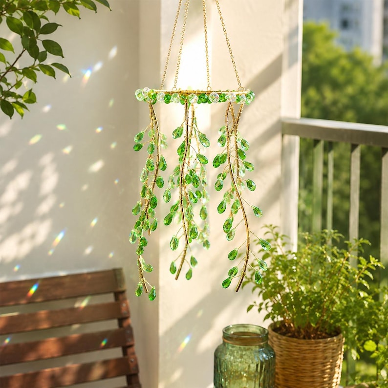 May include: A hanging mobile featuring a circular gold-toned frame suspended by three thin chains. Attached to the frame are multiple cascading strands of wire adorned with faceted, teardrop-shaped green glass crystals that catch the sunlight.