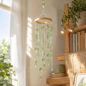 Può includere: Una giostrina sospesa con un cerchio in legno circolare appeso a catene argentate. Molteplici fili di perline trasparenti e verdi a forma di cuore pendono dal cerchio, creando un effetto a cascata intervallato da piccoli cristalli.