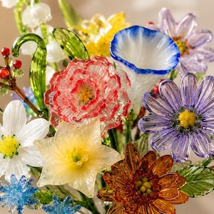 May include: A bouquet of handcrafted glass flowers featuring a red-tipped carnation, a blue-rimmed white bell-shaped flower, a purple daisy, a yellow daffodil, a white flower, a small blue flower, and green glass leaves with red berries.