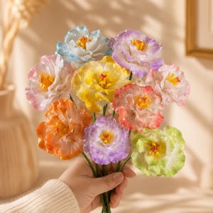 Op de afbeelding: Een boeket kunstbloemen met meerdere bloesems in diverse kleuren, waaronder blauw, geel, paars, oranje, roze en lichtgroen. Elke bloem heeft gegolfde bloemblaadjes en een felgeel hart, bevestigd aan dunne groene stelen.
