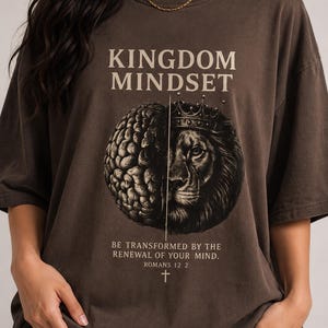 Könnte beinhalten: Ein braunes T-Shirt mit dem Aufdruck "KINGDOM MINDSET" oben. Darunter befindet sich eine Grafik eines Gehirns, das in zwei Hälften geteilt ist, mit einem Löwen mit Krone. Der Text "BE TRANSFORMED BY THE RENEWAL OF YOUR MIND. ROMANS 12:2" ist darunter gedruckt.