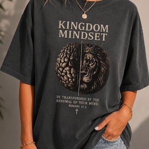 Op de afbeelding: Donkergrijs t-shirt met de tekst "KINGDOM MINDSET" boven een afbeelding van een hersenhelft en een leeuwenkop met een kroon. De tekst "BE TRANSFORMED BY THE RENEWAL OF YOUR MIND. ROMANS 12 2" staat eronder, met een kruis.