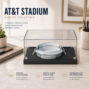 May include: A desktop collectible of AT&T Stadium, a detailed model inside a clear display case. The stadium model is gray and white, set on a black base. The text reads "AT&T STADIUM DESKTOP COLLECTIBLE".