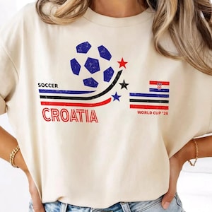 May include: A cream-colored t-shirt featuring a graphic with a blue soccer ball, three stars in red, black, and blue, and horizontal stripes. The text reads SOCCER CROATIA and WORLD CUP '26, accompanied by a small Croatian flag graphic.