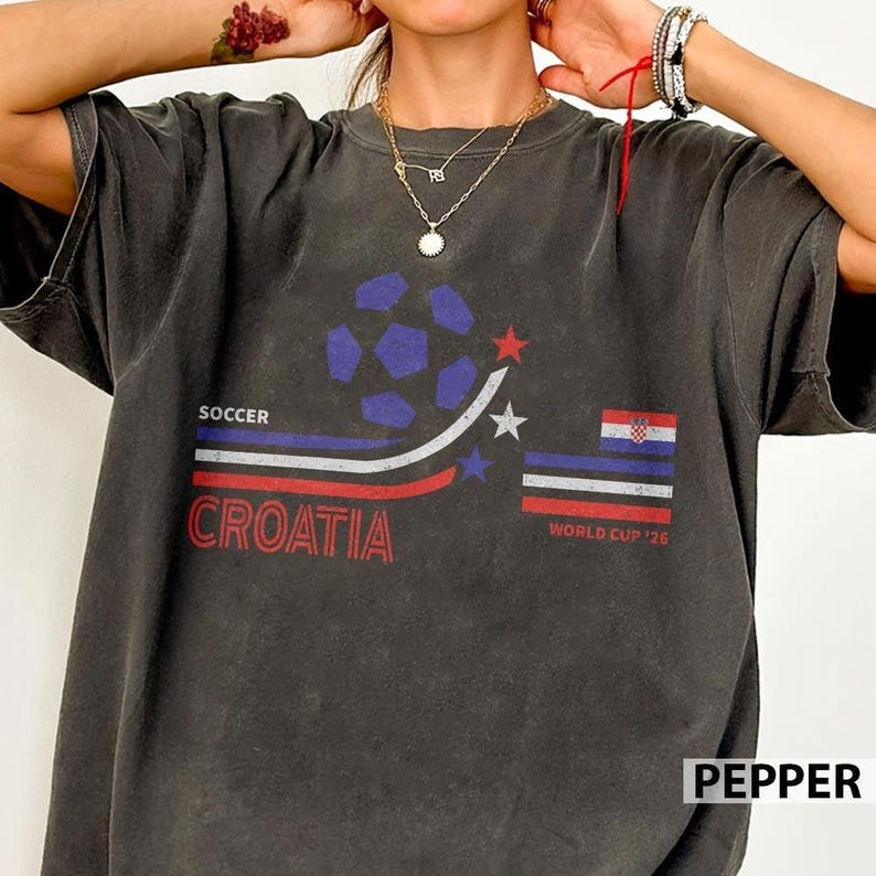 May include: A charcoal grey short-sleeve t-shirt featuring a graphic print with a blue soccer ball, red and blue stripes, and the text SOCCER CROATIA. The design includes a small Croatian flag, white stars, and the text WORLD CUP '26 in red.
