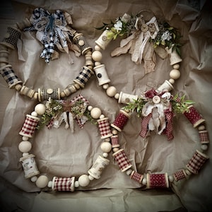 May include: Four circular wreaths constructed from wooden beads and vintage-style wooden spools wrapped in various fabrics, including black and white buffalo check, red gingham, and floral prints, accented with fabric bows, greenery, and buttons.