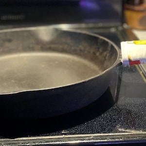 May include: A well-used, black cast iron skillet with a white and yellow fruit-patterned handle cover rests on a black stovetop. The skillet is round with a single handle and shows signs of wear. The handle cover adds a touch of color and protection.