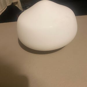 May include: A smooth, rounded, white glass lamp shade with a dome-like shape. The object features a matte, opaque finish that curves gently toward a central peak, creating a soft, organic silhouette that is uniform in color and texture throughout.