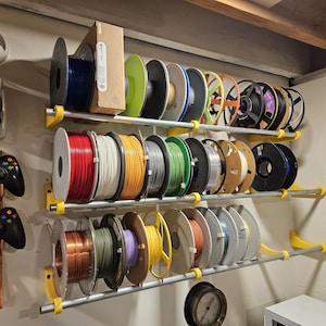 May include: A wall-mounted storage system with three shelves holding numerous spools of 3D printing filament in various colours. The shelves are supported by yellow brackets. Also visible are two black game controllers and a VR headset.