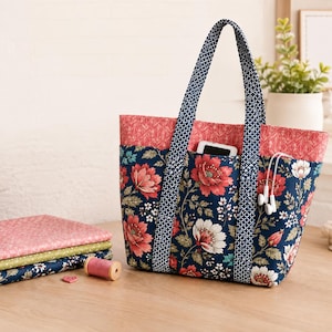 Peut inclure: Un sac fourre-tout en tissu matelassé avec un fond bleu marine orné de fleurs rouges et blanches, une bande supérieure rose corail et des anses à motifs bleu marine. Une pile de tissus pliés et une bobine de fil en bois sont posées à côté.