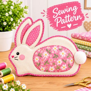 May include: A fabric bunny-shaped sewing pattern project featuring cream-colored fabric with a pink floral center panel, pink polka-dot ears, a white pom-pom tail, and a pink heart nose. The text Sewing Pattern is displayed in white.