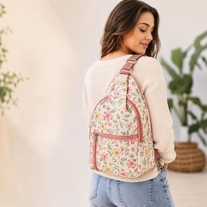 May include: A sling backpack featuring a cream-colored base with a multi-colored floral pattern of pink, yellow, and green flowers. The bag includes a front zippered pocket, pink trim, and an adjustable shoulder strap with metallic hardware.