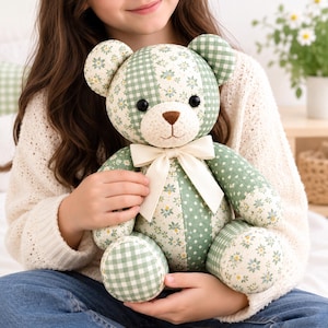 May include: A stuffed teddy bear crafted from patchwork fabric featuring alternating green and white gingham patterns and floral prints. The bear has black button eyes, a brown stitched nose, and a cream-coloured ribbon tied around its neck.