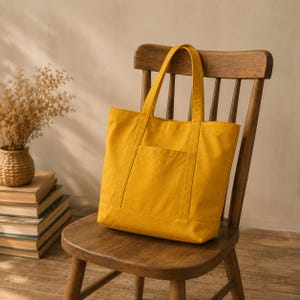 May include: A mustard yellow canvas tote bag featuring two sturdy shoulder straps and a single rectangular exterior pocket on the front. The bag has a structured rectangular shape with reinforced stitching along the seams and handles.