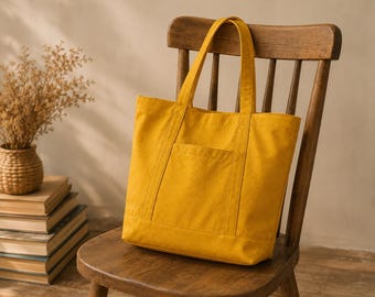 May include: A mustard yellow canvas tote bag featuring two sturdy shoulder straps and a single rectangular exterior pocket on the front. The bag has a structured rectangular shape with reinforced stitching along the seams and handles.