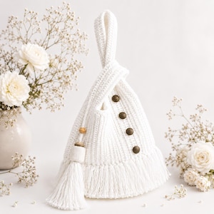 Puede incluir: Bolso blanco de crochet hecho a mano, con asa anudada y tres botones decorativos. El bolso presenta flecos en la parte inferior y una borla con cuentas de madera. Flores blancas en un jarrón completan la escena.
