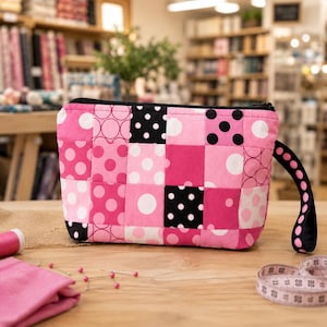 May include: A rectangular zipped pouch featuring a patchwork design of pink, white, and black fabric squares with various polka dot and circular patterns. The pouch includes a matching black wrist strap with white polka dots attached to the side.