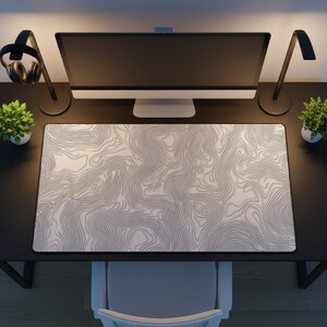 May include: A rectangular mousepad featuring a white and grey topographic map design. The mousepad is on a black desk, with a computer monitor, lamps, and small potted plants. The mousepad has a black border.
