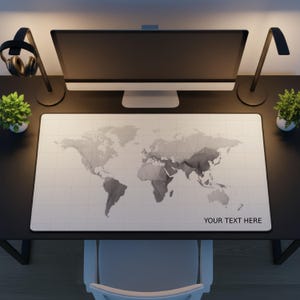 May include: A rectangular white desk mat with a grayscale world map design and a grid pattern. The words "YOUR TEXT HERE" are printed in black at the bottom right. The mat is on a black desk with a computer monitor, lamps, and small plants.