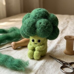 May include: A needle-felted broccoli figure with a textured dark green floret top and a light green stem body. The figure features two small black bead eyes and a simple stitched mouth, sitting upright with its arms resting on its knees.