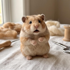 Könnte beinhalten: Eine nadelgefilzte Hamsterfigur mit texturiertem hellbraunem und cremefarbenem Wollfell, dunklen Perlenaugen und einer kleinen rosa Nase. Der Hamster steht aufrecht mit erhobenen Vorderpfoten und hat ein realistisches Aussehen sowie eine flauschige Textur.