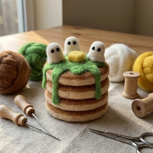 Puede incluir: Una pila de tres tortitas de lana fieltrada de color tostado, cubiertas con sirope verde, un trozo de mantequilla amarillo y tres pequeños fantasmas blancos con ojos de cuentas negras, rodeada de ovillos de lana, carretes y herramientas.