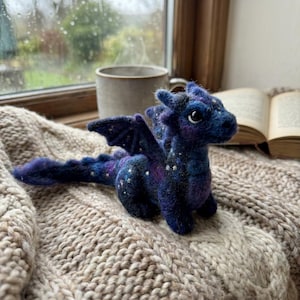 May include: A needle-felted dragon figurine in shades of deep blue, purple, and black, featuring small white star-like accents on its body, wings, and long tail. The creature has large dark eyes and small horns, crafted with a textured, fuzzy wool finish.