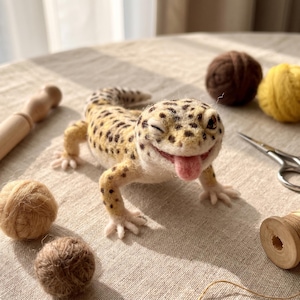 May include: A needle-felted leopard gecko figurine with yellow and brown spotted skin, a winking eye, and a small pink tongue sticking out. The gecko is positioned among various craft supplies including yarn balls, a wooden spool, and scissors.