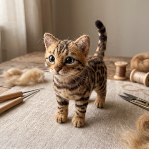 May include: A needle-felted kitten figurine with a tan and brown tabby coat featuring dark stripes and spots. The small cat has large black eyes, a pink nose, long white whiskers, and a tail held upright, standing on a textured fabric surface.