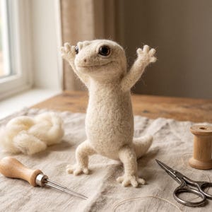 May include: A needle-felted cream-colored gecko figurine standing upright with its front limbs raised. The gecko has large dark eyes and a textured wool surface. It is positioned alongside a felting tool, a wooden spool, and loose wool.