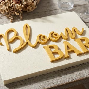 Op de afbeelding: Een decoratief bord met de woorden "Bloom BAR" in gele, cursieve letters. Het bord staat op een wit, getextureerd oppervlak, met een houten tafel op de achtergrond. Er zijn ook gedroogde bloemen en glazen te zien.