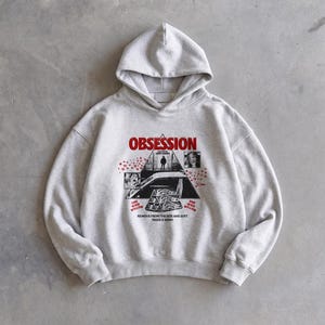 Puede incluir: Una sudadera con capucha gris claro que presenta un estampado gráfico en el pecho. El diseño incluye la palabra OBSESSION en rojo, un marco triangular central con una silueta, pequeñas imágenes insertadas, iconos de corazones rojos y el texto REMOVE FROM THE BOX AND JUST MAKE A WISH.