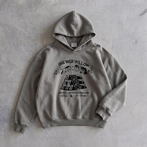 May include: A gray hoodie with a drawstring hood and a kangaroo pocket. The front features a black graphic with the text "ONE WISH WILLOW" and other illustrations. The hoodie is made of a soft, comfortable material.