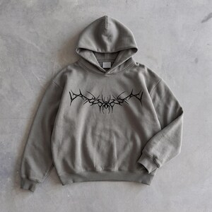 Puede incluir: Una sudadera con capucha gris con un dise&ntilde;o gr&aacute;fico negro y puntiagudo en el pecho. La sudadera tiene capucha y mangas largas. El dise&ntilde;o es un patr&oacute;n sim&eacute;trico estilizado. La sudadera est&aacute; hecha de un material suave.
