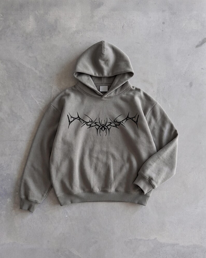 Puede incluir: Una sudadera con capucha gris con un dise&ntilde;o gr&aacute;fico negro y puntiagudo en el pecho. La sudadera tiene capucha y mangas largas. El dise&ntilde;o es un patr&oacute;n sim&eacute;trico estilizado. La sudadera est&aacute; hecha de un material suave.