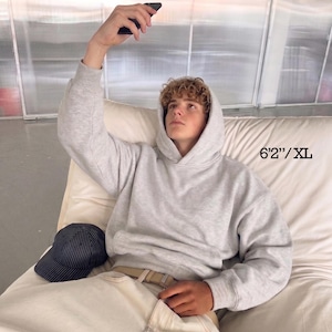 May include: A light gray hoodie with a drawstring hood and a front pocket. The model is wearing light-colored pants and a baseball cap. The text on the image reads "6'2"/XL".