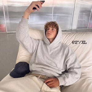 Puede incluir: Una sudadera con capucha gris claro con cord&oacute;n y bolsillo delantero. El modelo lleva pantalones de color claro y una gorra de b&eacute;isbol. El texto de la imagen dice "6'2"/XL".