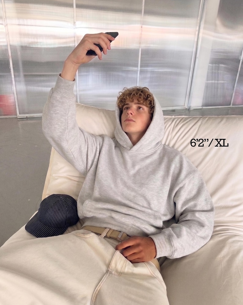 Puede incluir: Una sudadera con capucha gris claro con cord&oacute;n y bolsillo delantero. El modelo lleva pantalones de color claro y una gorra de b&eacute;isbol. El texto de la imagen dice "6'2"/XL".