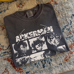 May include: A charcoal gray crew neck t-shirt featuring a white graphic print of three manga-style character portraits arranged in panels. The text ACKERMAN is printed in a stylized font above the panels. The shirt has a Comfort Colors brand label.