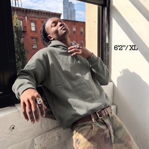 May include: A sage green hoodie with a drawstring hood and long sleeves. The model is wearing a brown belt and camouflage pants. The text "6'2"/XL" is visible in the image.
