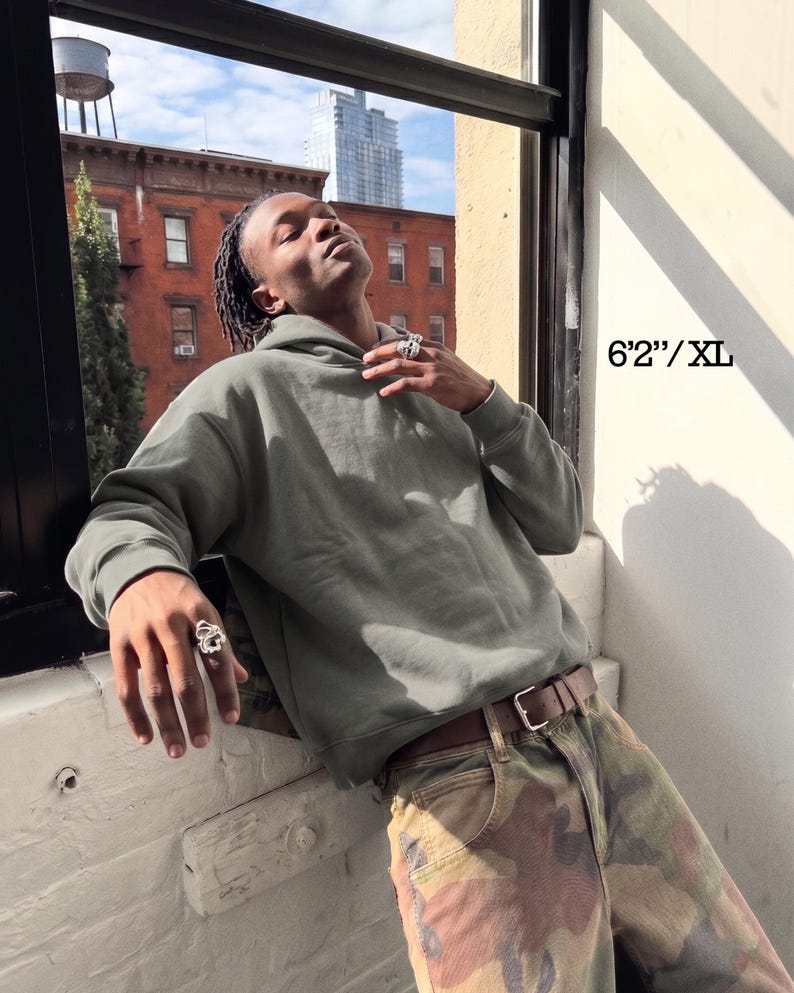 May include: A sage green hoodie with a drawstring hood and long sleeves. The model is wearing a brown belt and camouflage pants. The text "6'2"/XL" is visible in the image.