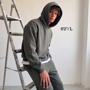 May include: A dark green hoodie and matching sweatpants. The model is wearing a white undershirt. The text "6'2"/L" is visible in the image. The person is leaning against a metal ladder.