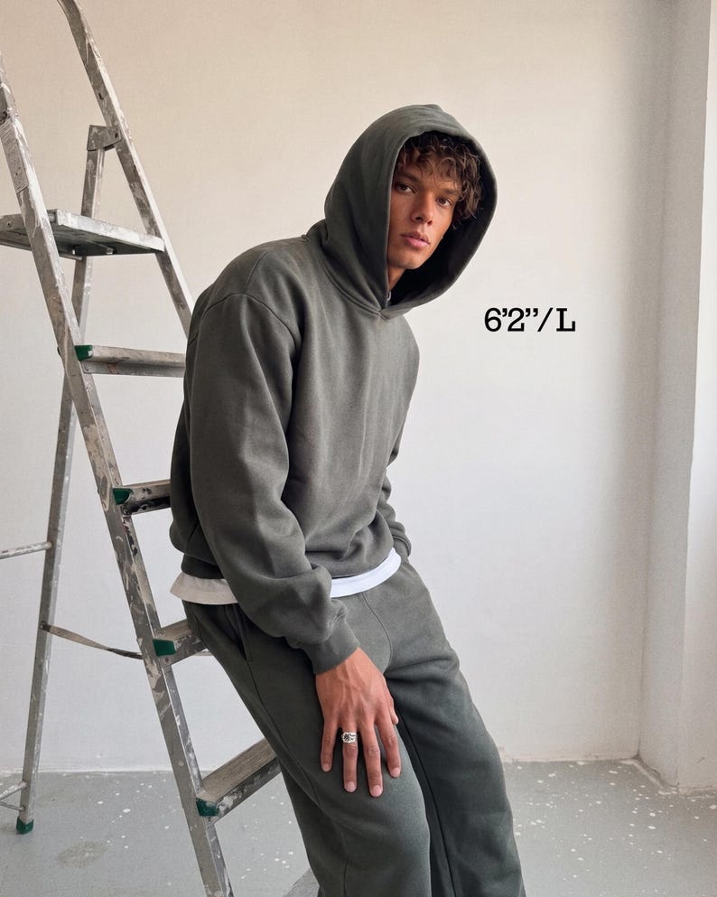May include: A dark green hoodie and matching sweatpants. The model is wearing a white undershirt. The text "6'2"/L" is visible in the image. The person is leaning against a metal ladder.