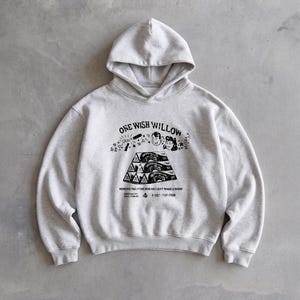 May include: A light gray hoodie with a hood and long sleeves. The front features a black graphic with the text "ONE WISH WILLOW" and other text and illustrations. The hoodie is made of a soft material.