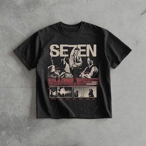 Op de afbeelding: Zwart t-shirt met een grafisch ontwerp uit de film "Se7en". Het ontwerp bevat de titel van de film in het wit, afbeeldingen van de acteurs en scènes uit de film. De tekst "film by David Fincher" is ook opgenomen.