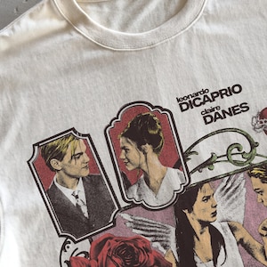 Pu&ograve; includere: Una t-shirt color crema con una stampa grafica di Leonardo DiCaprio e Claire Danes. Il design include due ritratti incorniciati, il testo "leonardo DICAPRIO claire DANES", una grande rosa rossa e l'illustrazione di una figura angelica.
