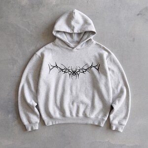 Puede incluir: Sudadera con capucha gris claro con un dise&ntilde;o gr&aacute;fico negro en el pecho. El gr&aacute;fico es un dise&ntilde;o estilizado y sim&eacute;trico con elementos puntiagudos y una forma central similar a una ara&ntilde;a. La sudadera tiene capucha y mangas largas.