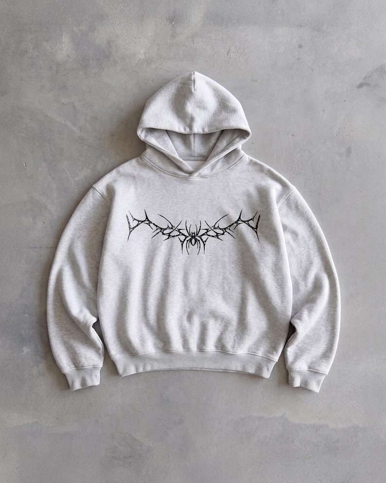 Puede incluir: Sudadera con capucha gris claro con un dise&ntilde;o gr&aacute;fico negro en el pecho. El gr&aacute;fico es un dise&ntilde;o estilizado y sim&eacute;trico con elementos puntiagudos y una forma central similar a una ara&ntilde;a. La sudadera tiene capucha y mangas largas.