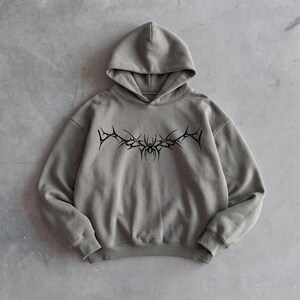 Puede incluir: Una sudadera con capucha gris con un dise&ntilde;o gr&aacute;fico negro en el pecho. El dise&ntilde;o parece ser un emblema estilizado y puntiagudo. La sudadera tiene capucha y mangas largas. La tela parece ser un material suave y de peso medio.
